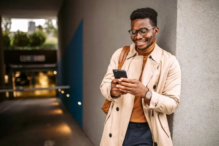 Smiling man in trench coat scrolling on phone in urban setting.