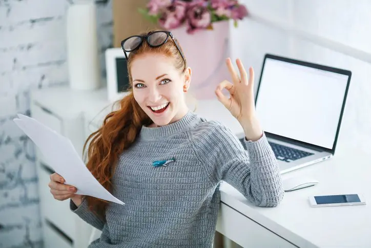 Young woman showing OK sign while holding papers at desk.