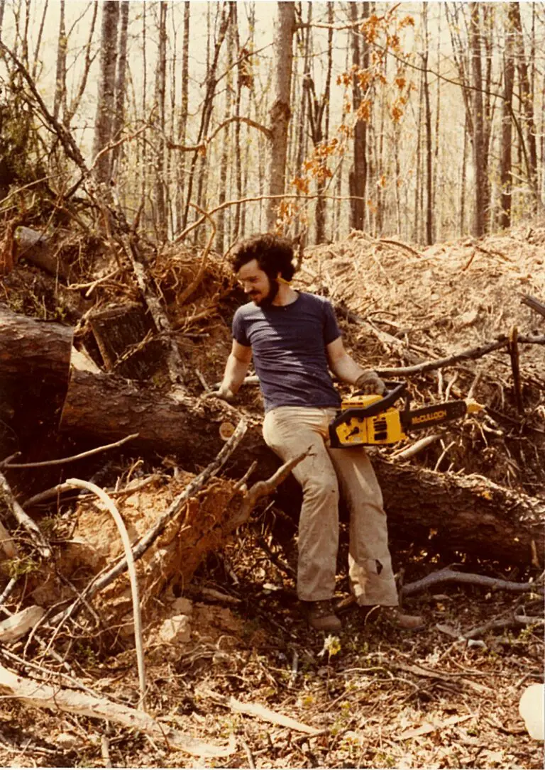 Man sitting on a fallen tree holding a chainsaw in a forest.